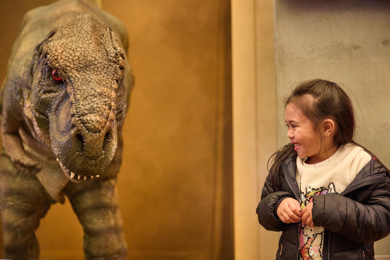 A young girl stands next to a T-Rex and look scared but excited.