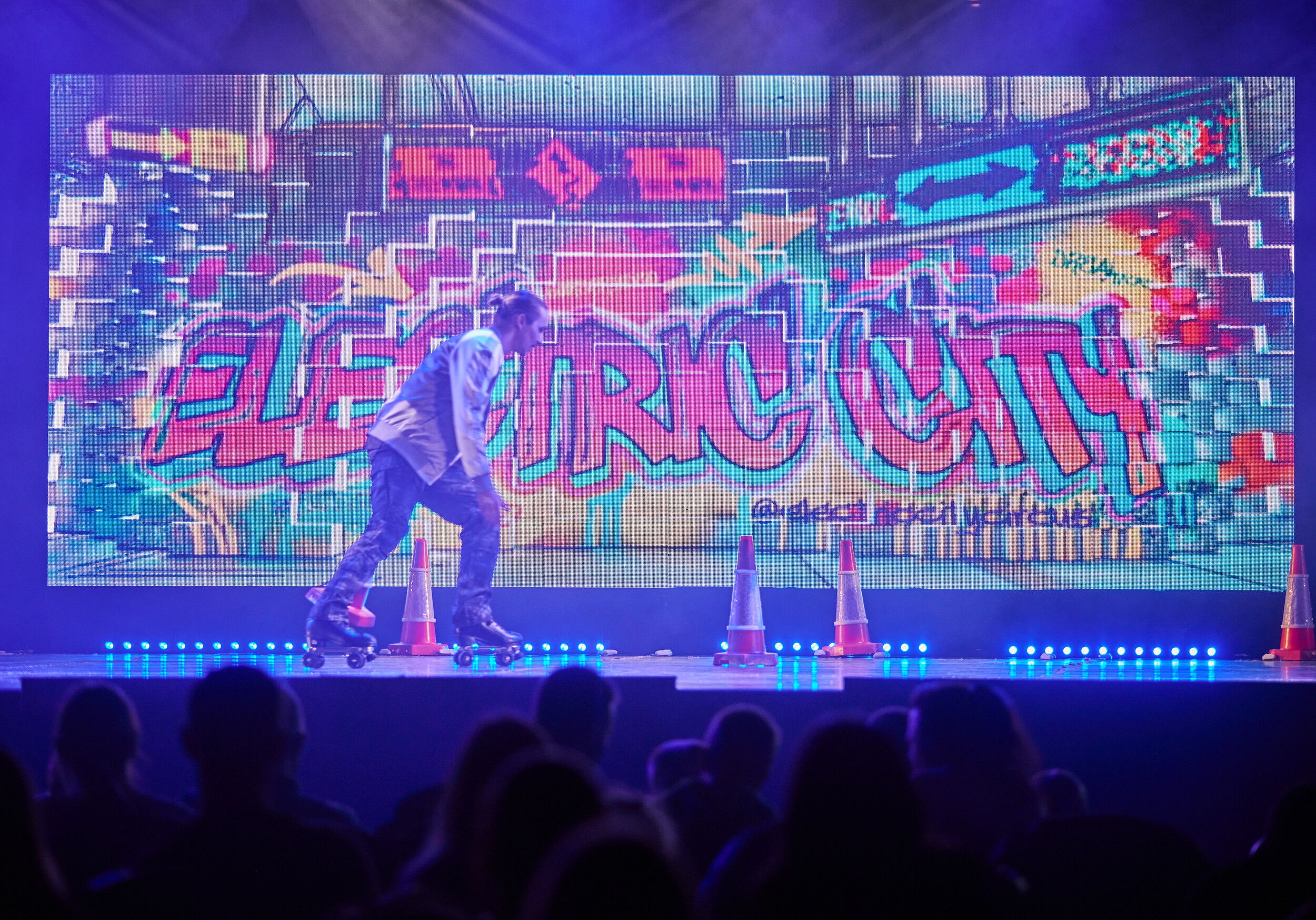 A man with roller skates performing on stage, at the background is a colourful sign saying "ELECTRIC CITY".
