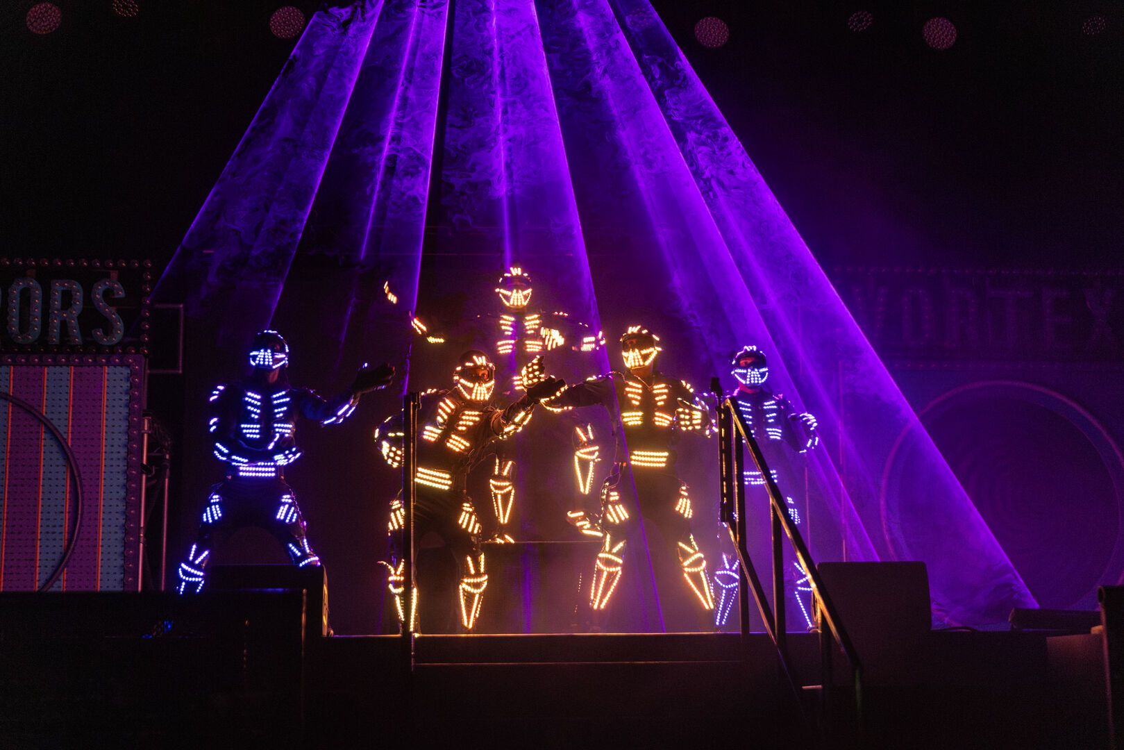 Five Electric Wonderland performers stand on stage in bright neon outfits, which are lit up in red and blue.