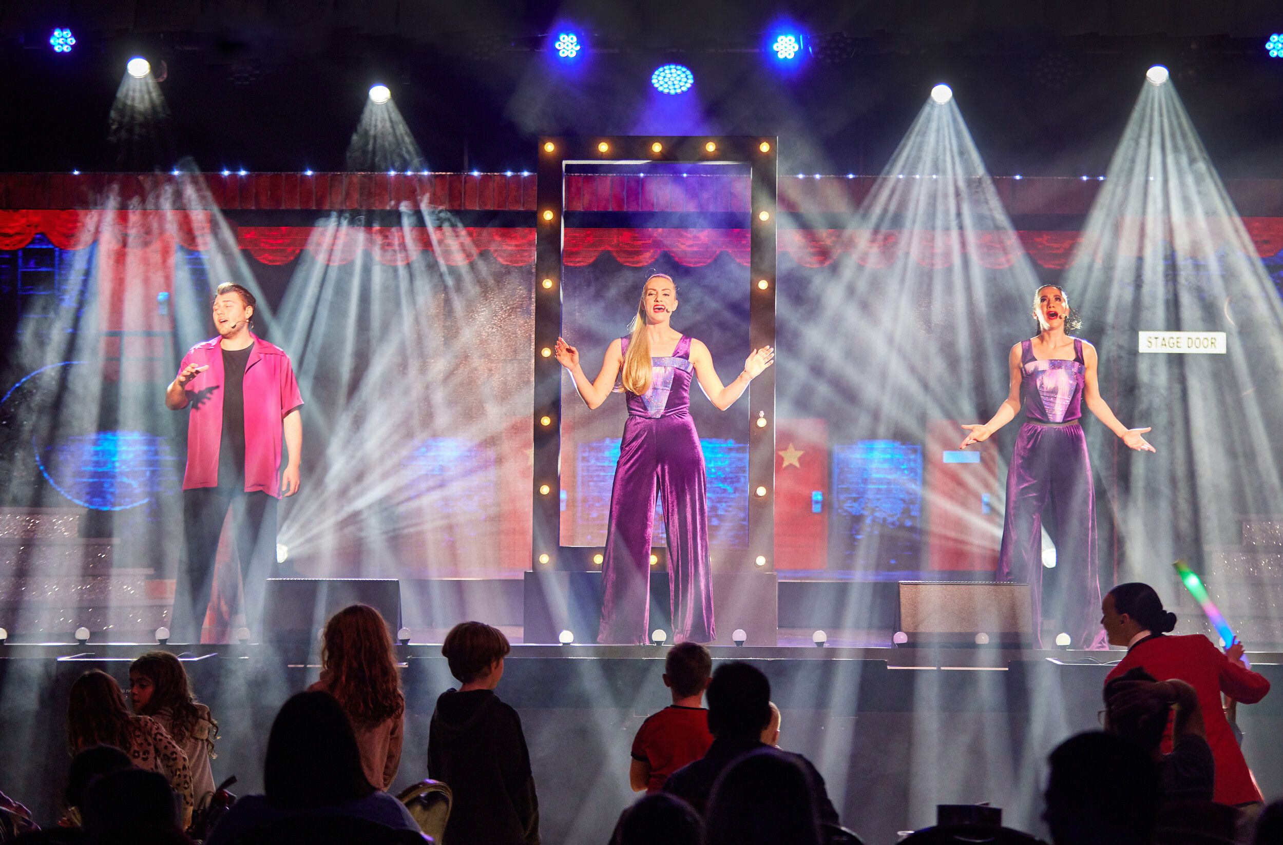A man in a red shirt and two women in purple dresses singing on stage under some white lights.