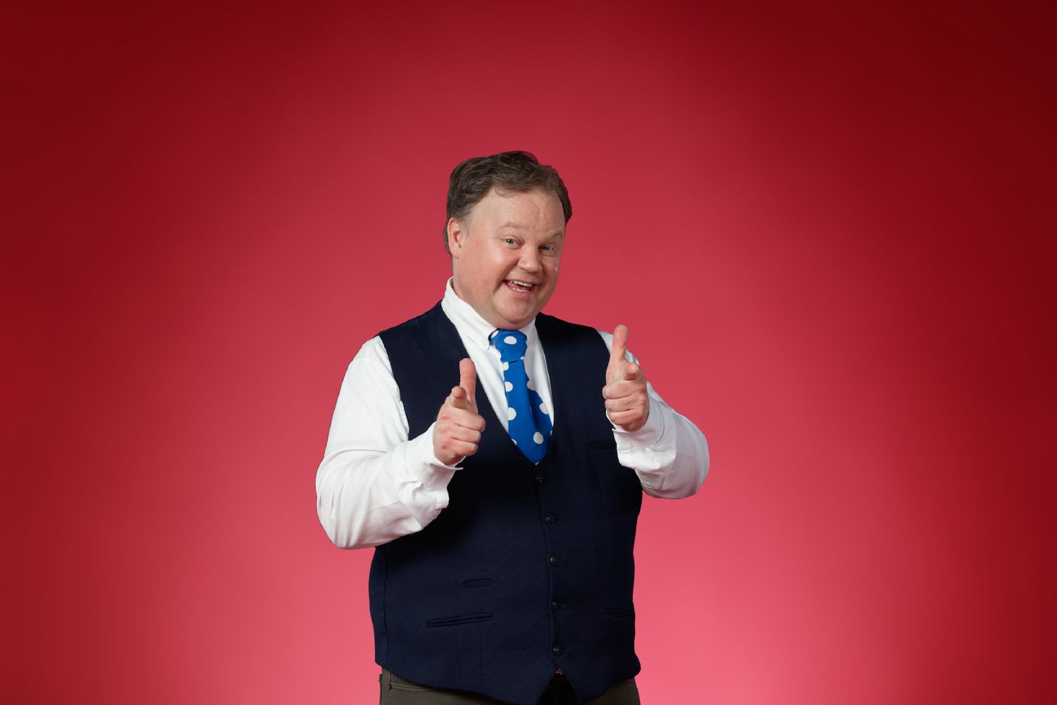 Justin Fletcher wearing a blue vest and tie while pointing at smiling at the camera.