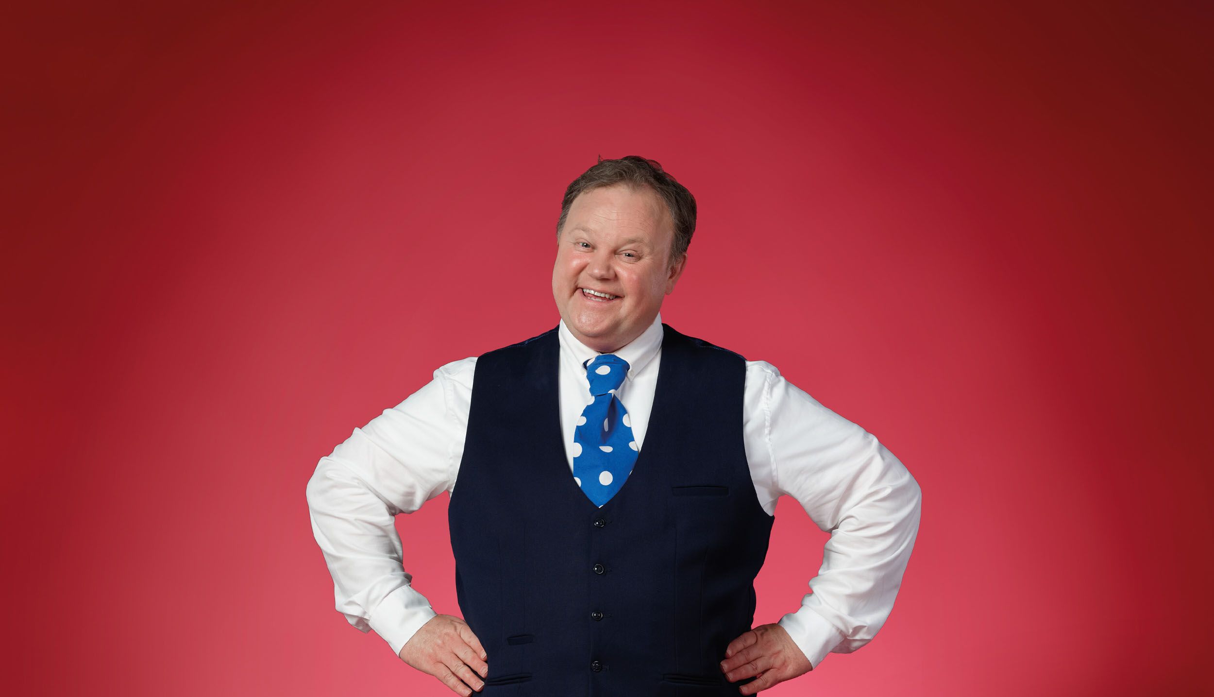 Justin Fletcher wearing a blue vest and tie while smiling at the camera.