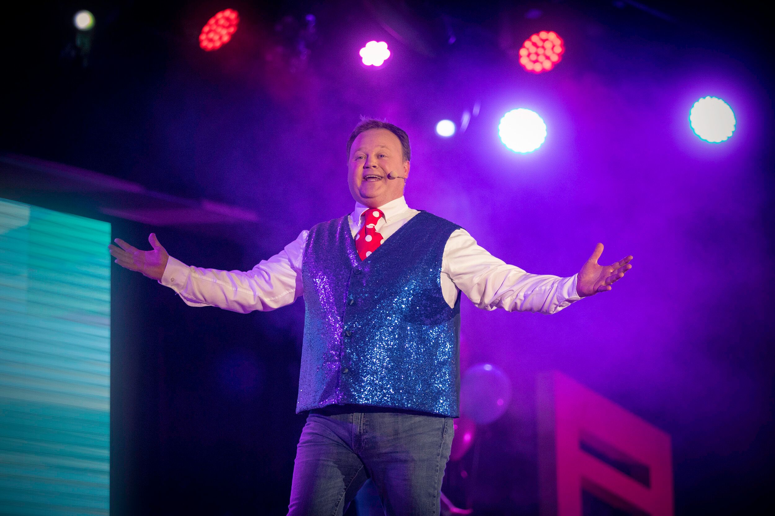 Justin Fletcher wearing a blue vest and red tie while performing on stage.