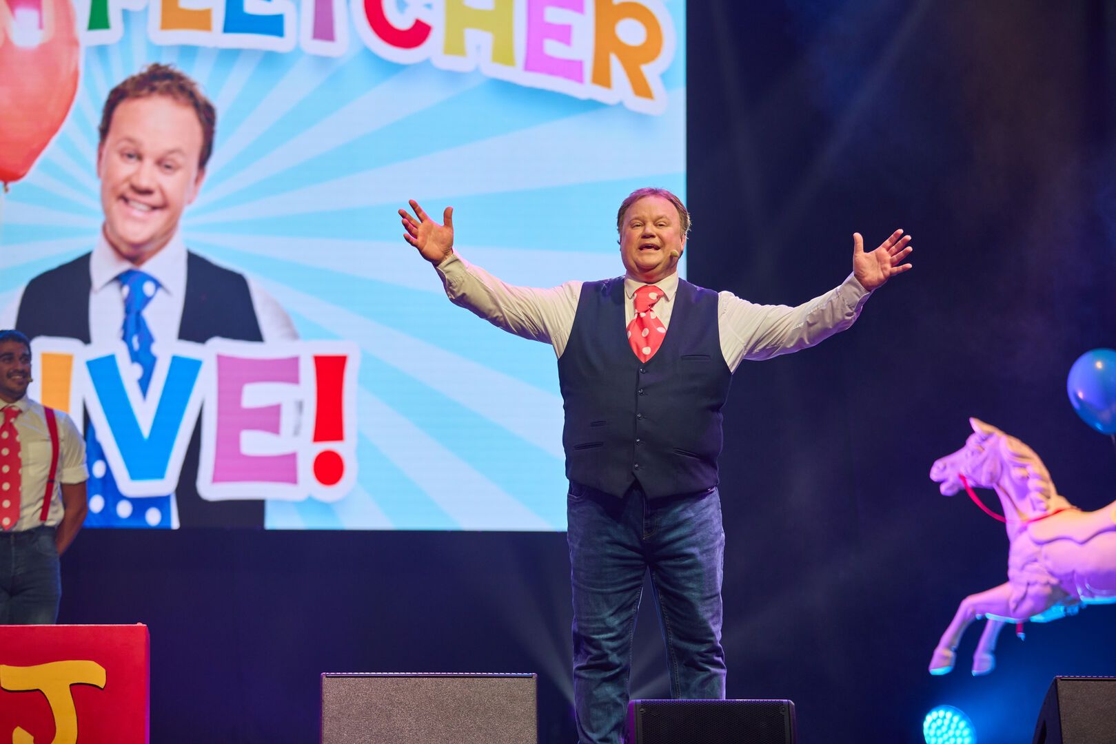 A man (Justin Fletcher) standing on a stage with his hand up in the air