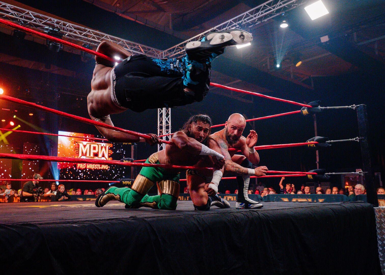 Two wrestlers are prone over the ropes. Another wrestler swings around, ready to tackle them both during a Maximum Pro Wrestling performance.