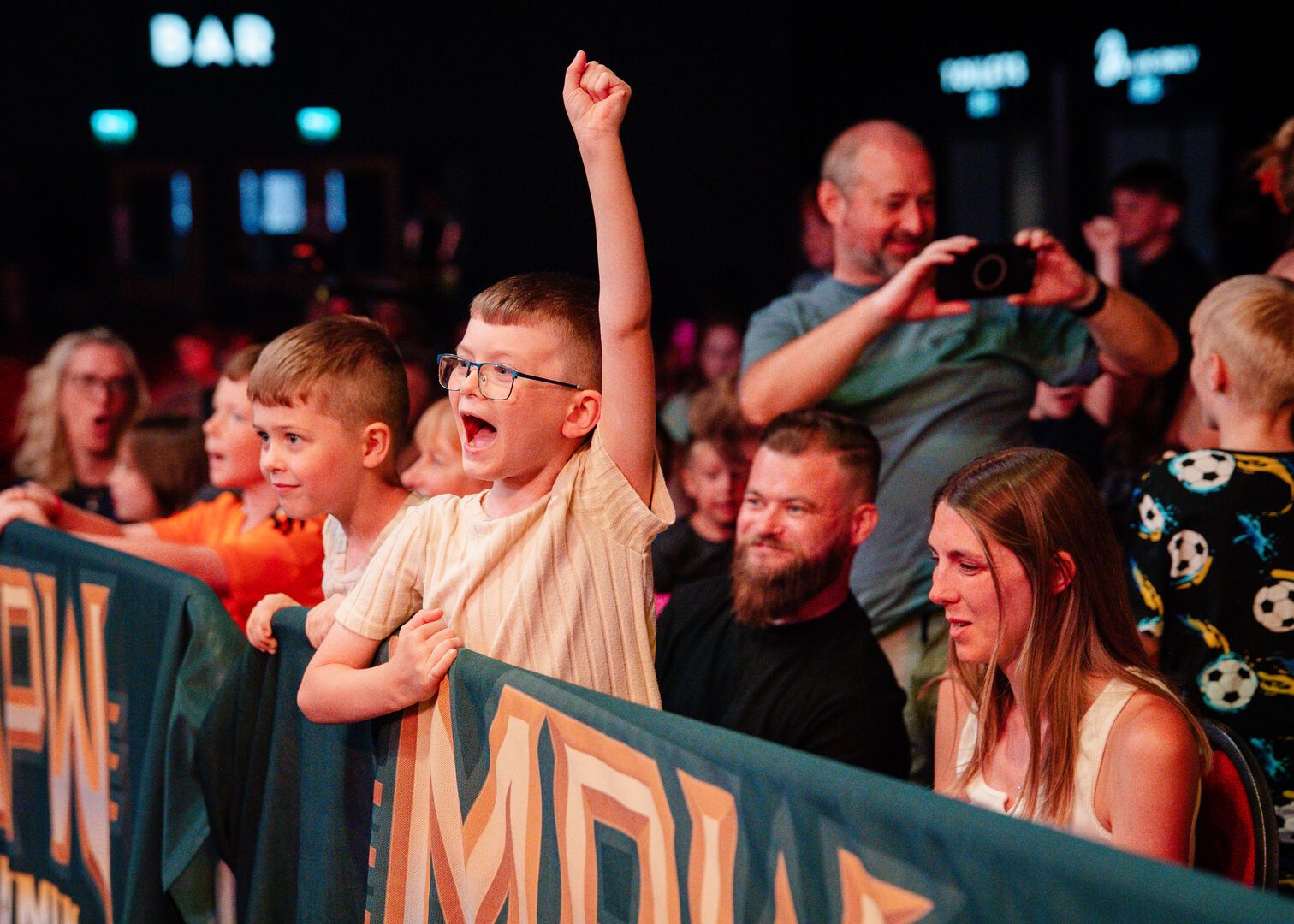 A photo of the audience during a Maximum Pro Wrestling show. A young boy stands at the front, with his hand in the air in excitement.