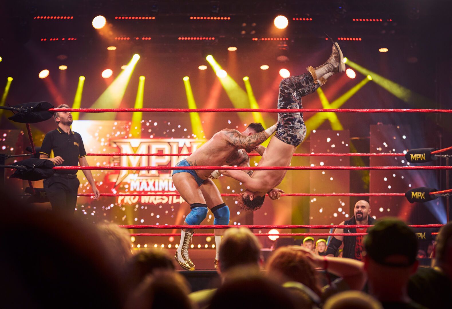 Two wrestlers fight in the ring as yellow and red stage-lighting frames them.