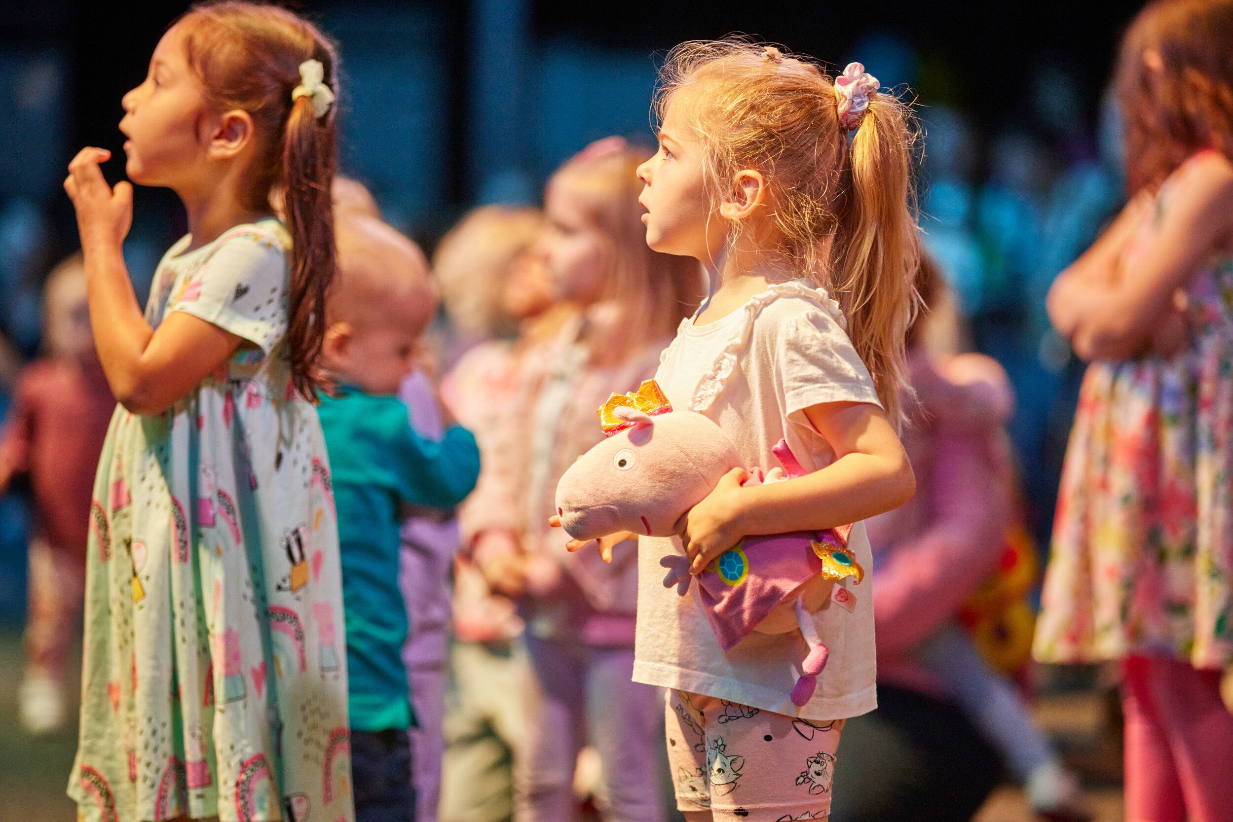 A little girl holding a Peppa Pig stuffed toy watching an event along with other kids.