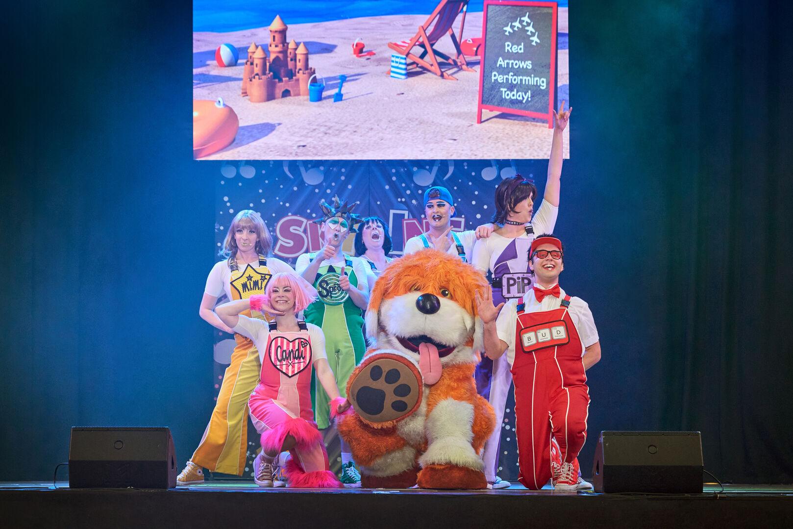The Skyline Gang cast huddle together on stage sheltering under umbrellas alongside their large orange dog mascot, with a giant binoculars graphic displayed on the screen backdrop above and the Skyline Gang logo visible behind them.