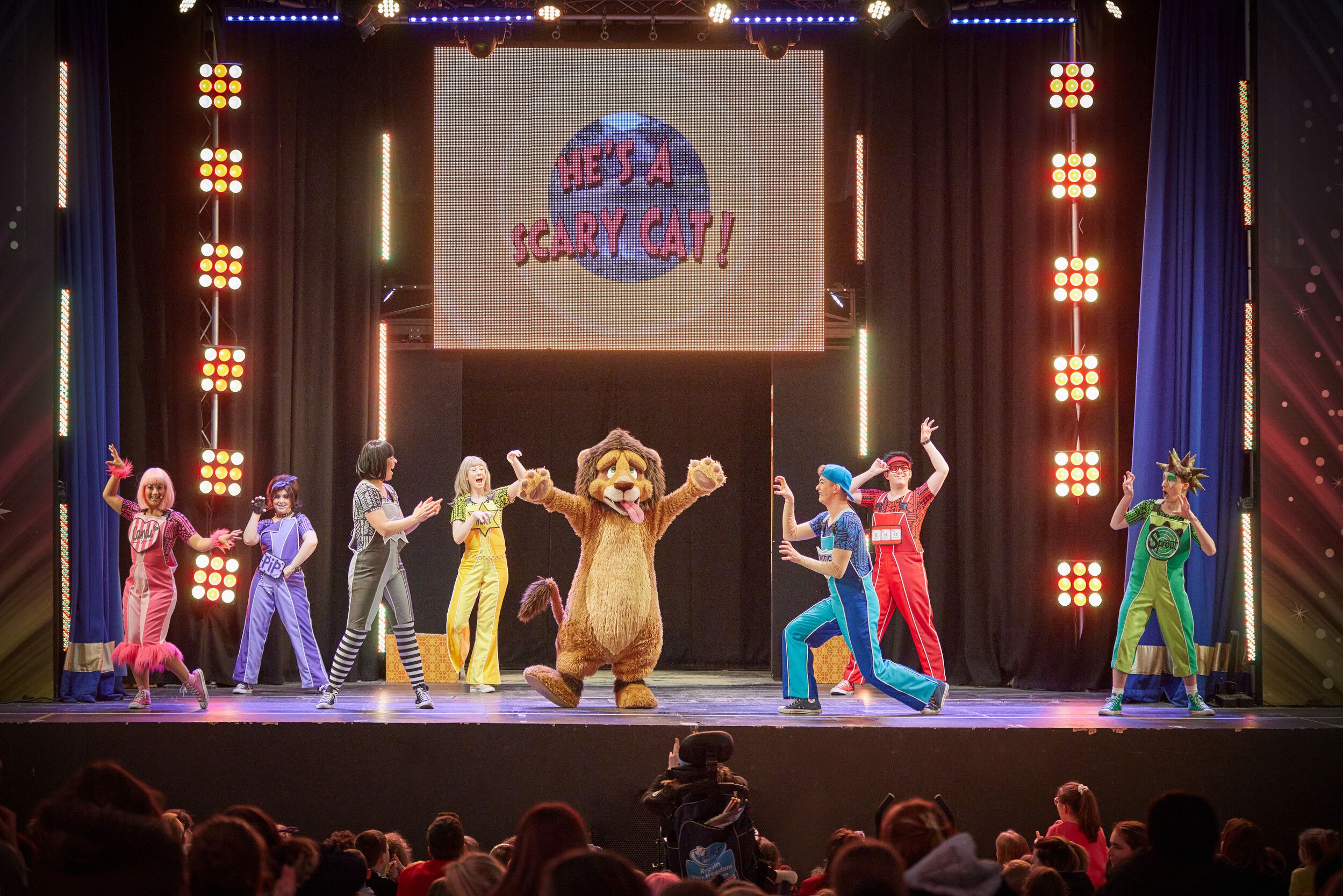 Skyline Gang members wearing colourful outfits while performing with a lion mascot on stage.