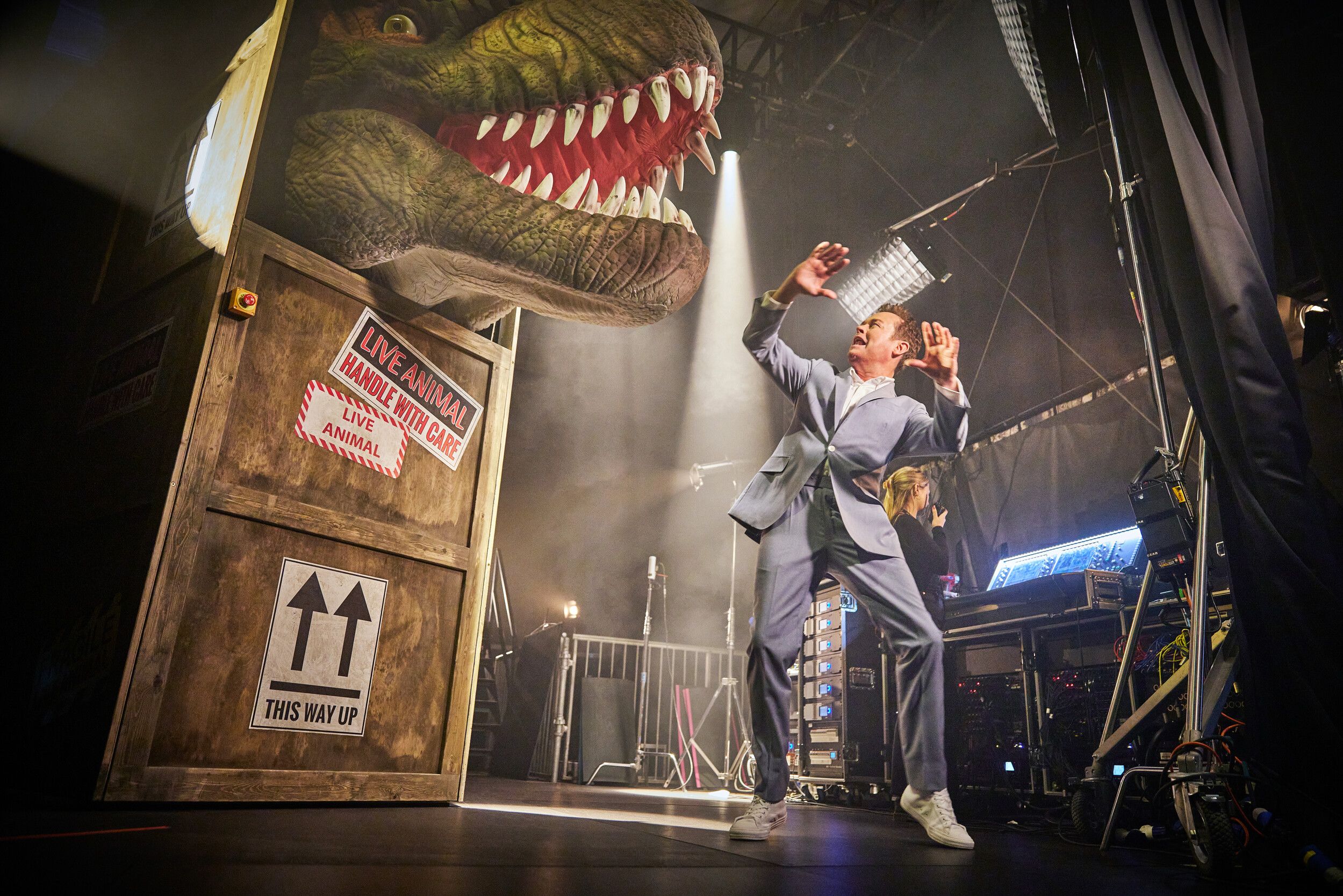 Stephen Mulhern wearing a grey suit doing shocked pose at a life-size T-Rex.