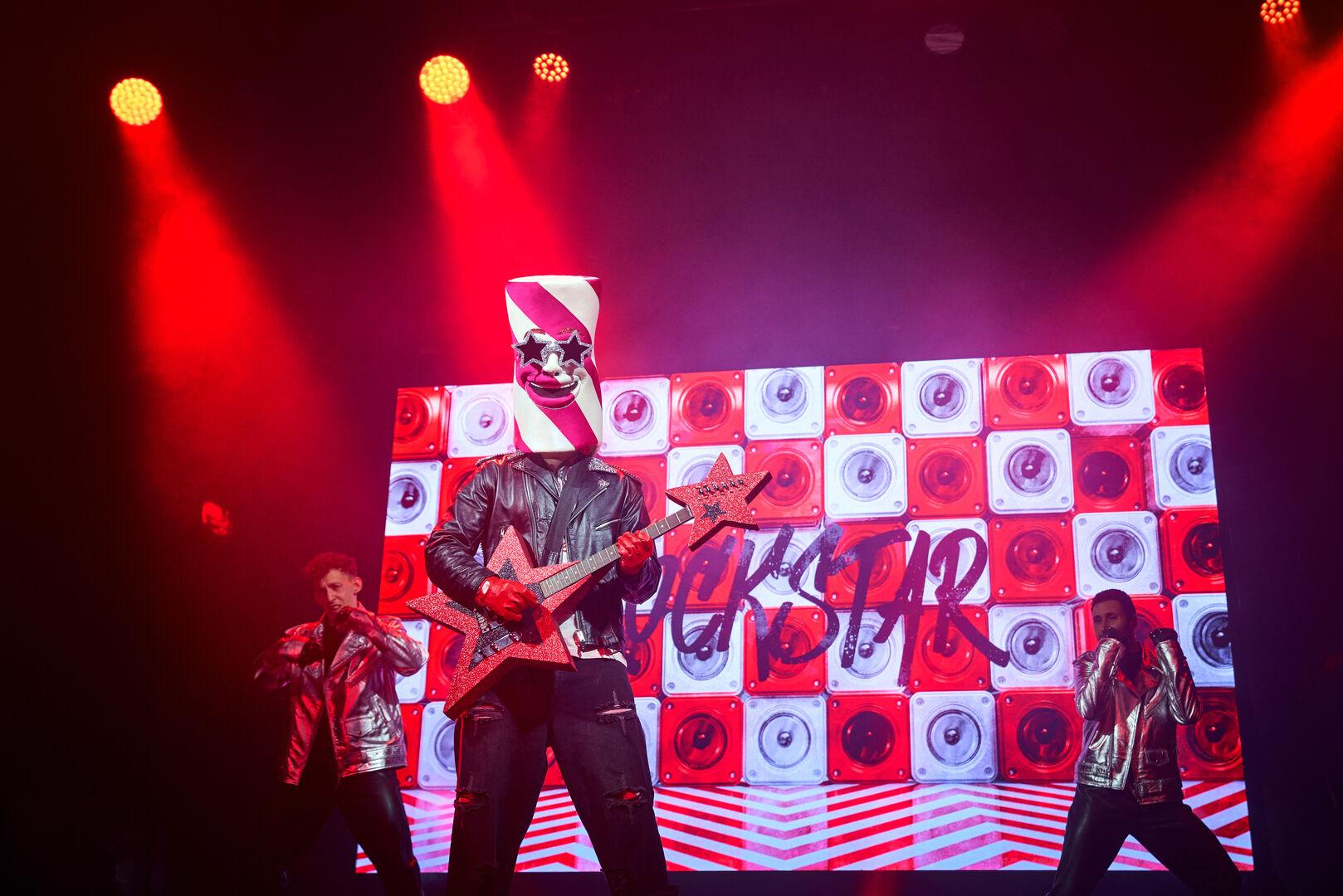 A wide shot of Rock-Star on stage playing a guitar, against a checkerboard backdrop and red lighting. 