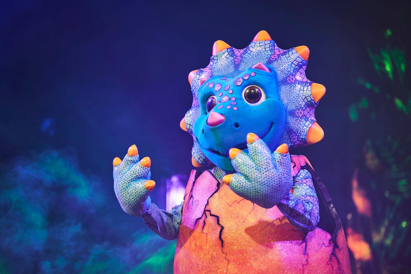 A colourful dinosaur costume character from The Masked Singer, featuring a blue scaly head with orange spikes, large expressive eyes, pink spots, and a friendly smile. The character wears a cracked egg shell body in orange and pink hues, with clawed gloved hands raised in a playful pose. The background is lit with atmospheric blue and green stage lighting with hints of purple crystal effects.