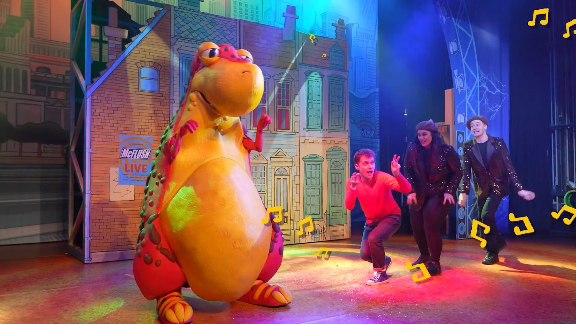A still taken from the stage show of the dinosaur that pooped. The Red, yellow and green dinosaur stands to the left of the stage, while three people are dancing on stage in a line, on the right.