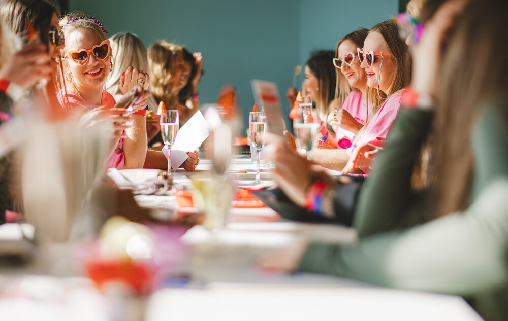 A group of people on a Big Weekender Bottomless Brunch, some are wearing heart shaped sun glasses while sitting around a dining table with wine glasses.