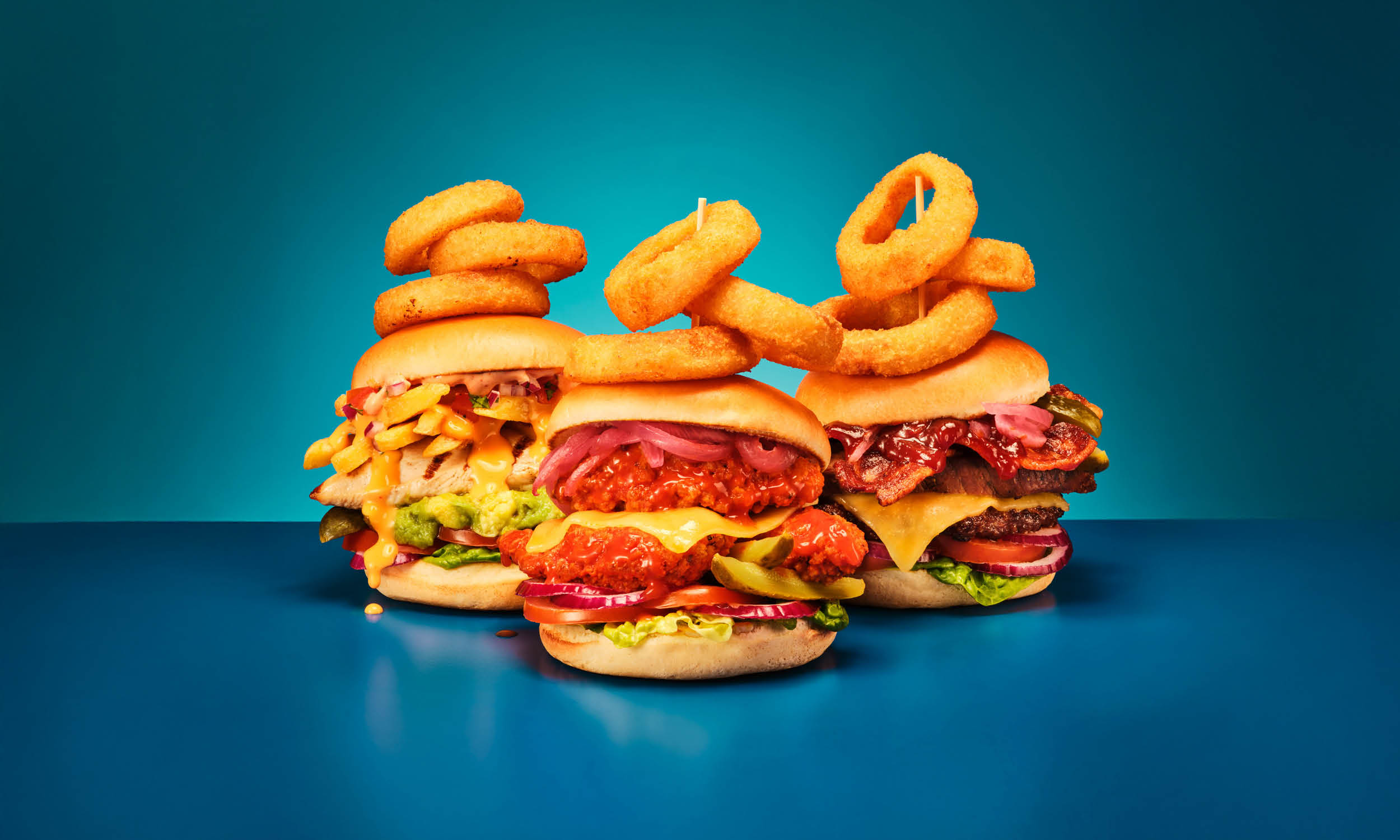 The Diner signature burgers. Three loaded burgers topped with stacks of onion rings on cocktail sticks against a graduated teal and blue background. Fillings include a grilled chicken burger with cheesy fries and guacamole, a buffalo chicken burger with pickled red onion, and a beef and bacon cheeseburger with barbecue sauce, lettuce and tomato.