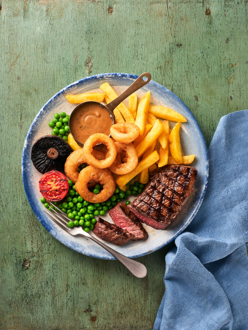 A plate of steak and chips, served with onion rings, peas, tomato, mushroom and baked beans.